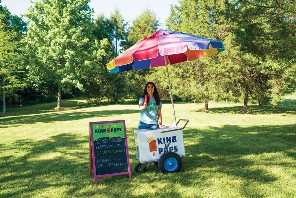 Woman selling colorful popsicles from a cart under a rainbow umbrella in a grassy park in Atlanta, Georgia.
