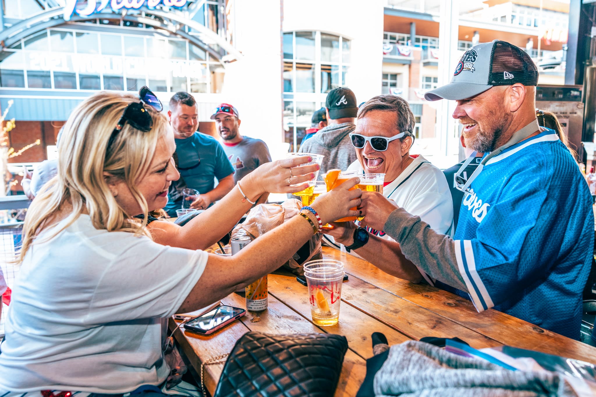 Three people cheerfully clink beer glasses at a crowded outdoor bar in Atlanta, Georgia.