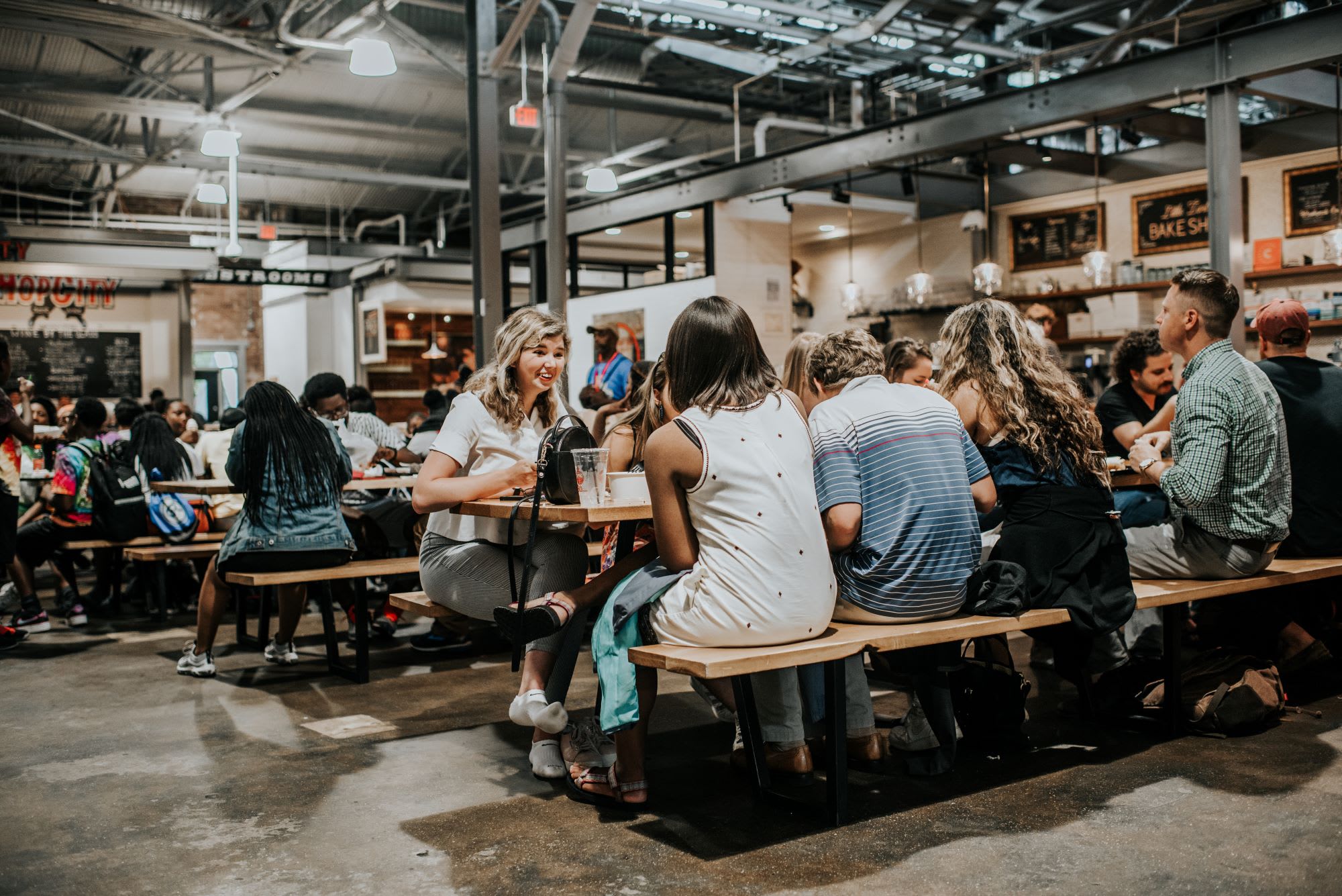 A diverse group of people enjoys a meal together at a long wooden table in a bustling indoor food court in Atlanta, Georgia.