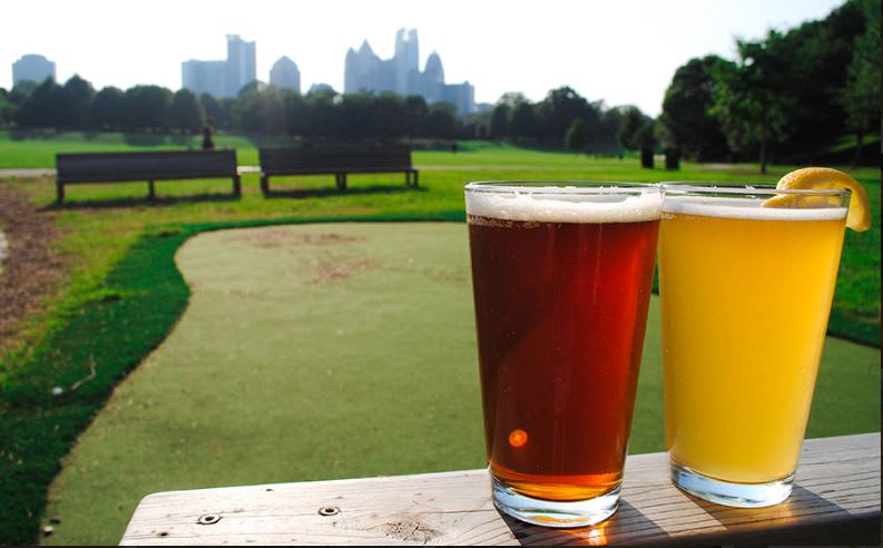 Two glasses of beer on a wooden railing overlook a green golf course with a city skyline in the distance in Atlanta, Georgia.