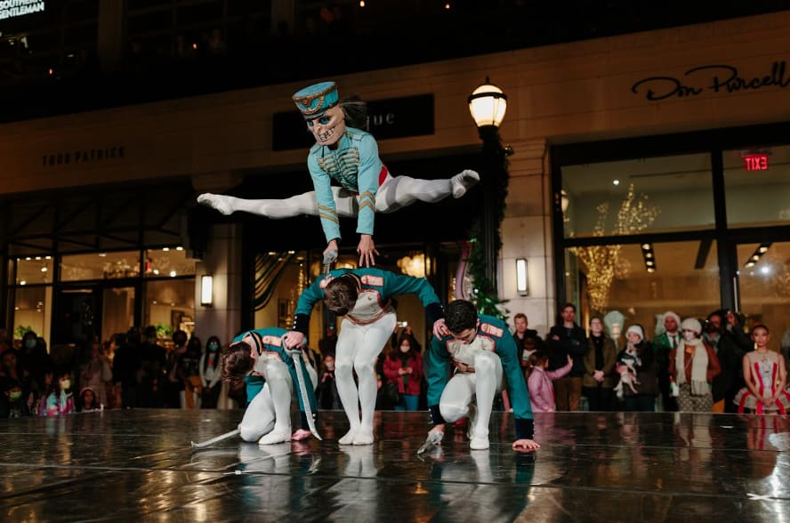 A ballet dancer in a nutcracker costume leaps over four kneeling dancers on a reflective stage in Atlanta, Georgia.