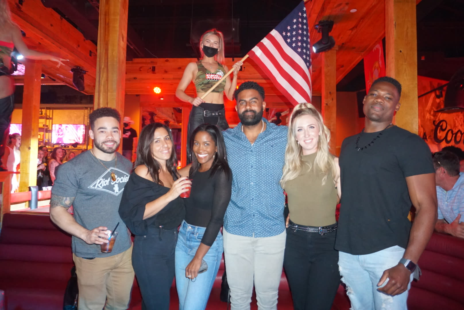 A diverse group of seven friends poses together at a lively bar or club, smiling and holding drinks in Atlanta, Georgia.