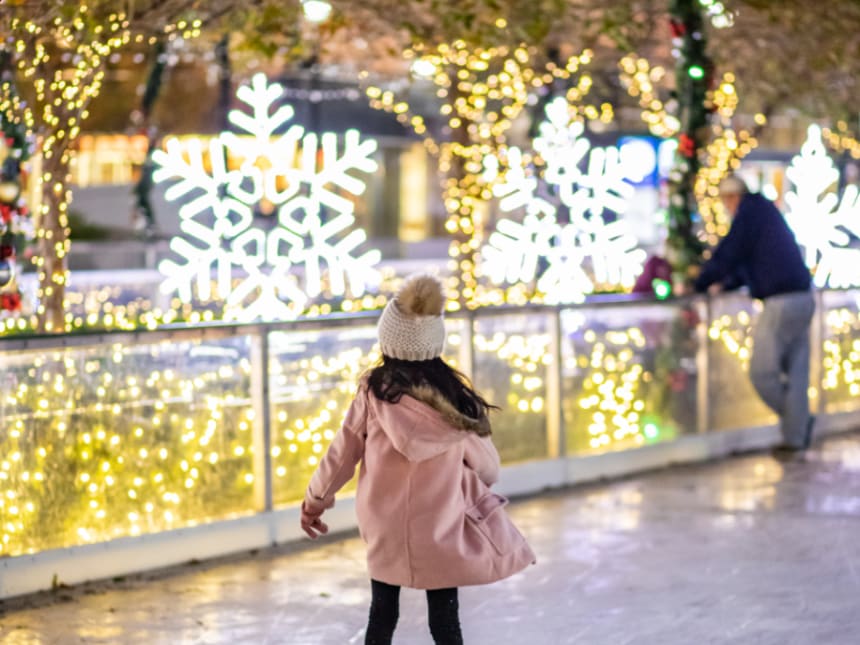 A young girl in a pink coat skates on an ice rink surrounded by festive lights and snowflake decorations in Atlanta, Georgia.