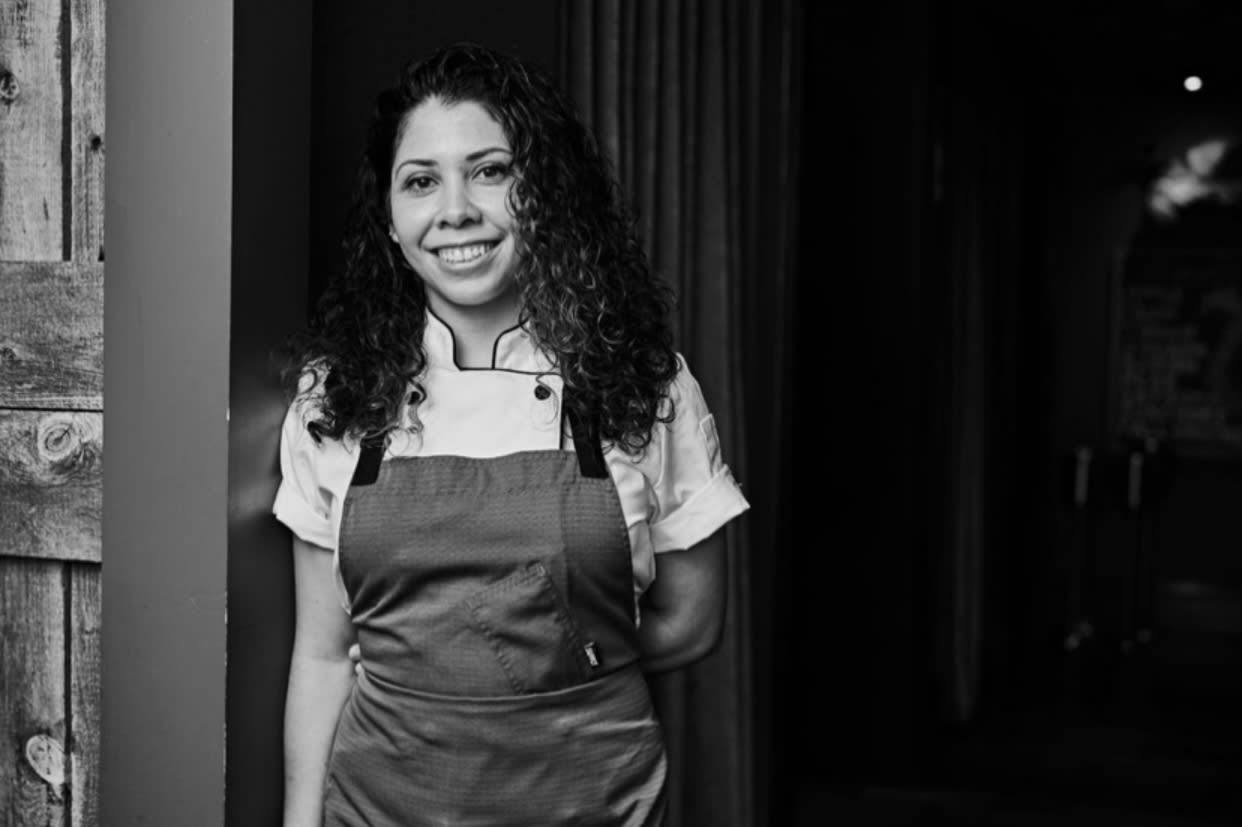 A smiling chef wearing a white shirt and apron stands by a wooden wall in Atlanta, Georgia.
