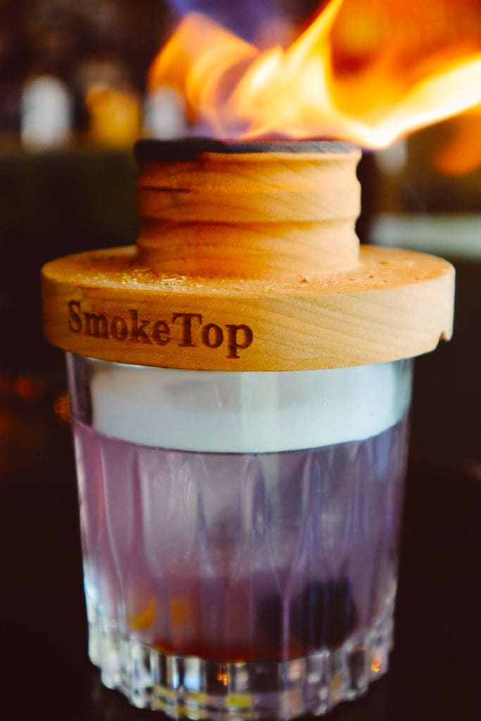 A glass filled with purple liquid and ice, topped with a wooden lid labeled 'SmokeTop', has a flame burning on top in Atlanta, Georgia.