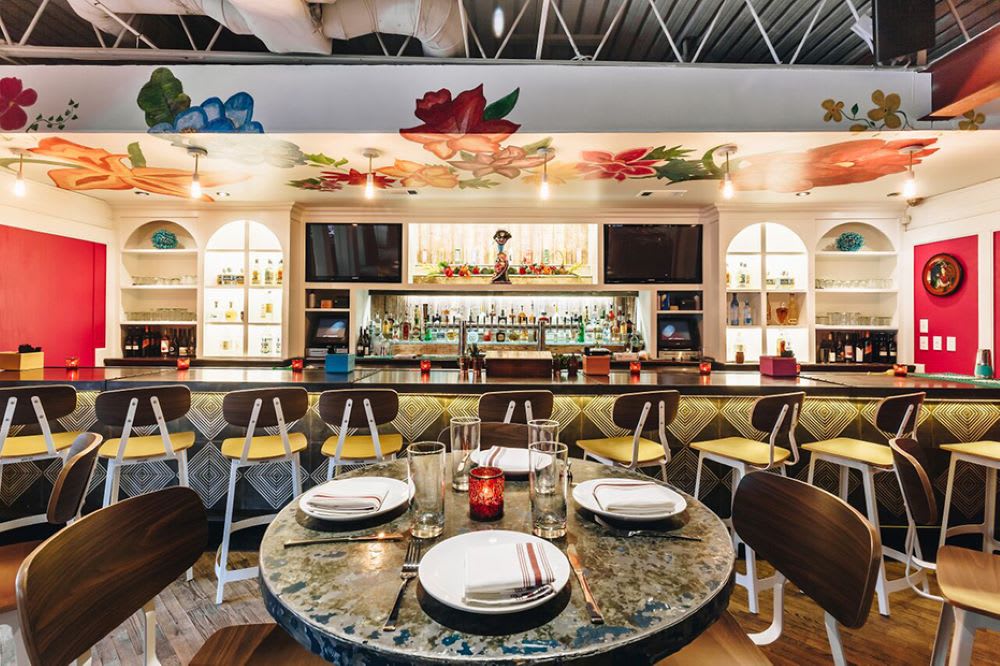 A vibrant restaurant interior with colorful floral ceiling murals, round tables, and a well-stocked bar in Atlanta, Georgia.