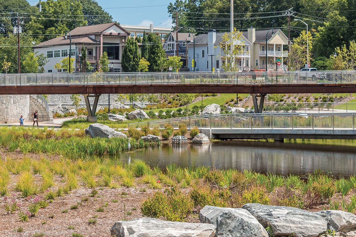 A pedestrian bridge spans a pond in a landscaped park with rocks and native plants in Atlanta, Georgia.