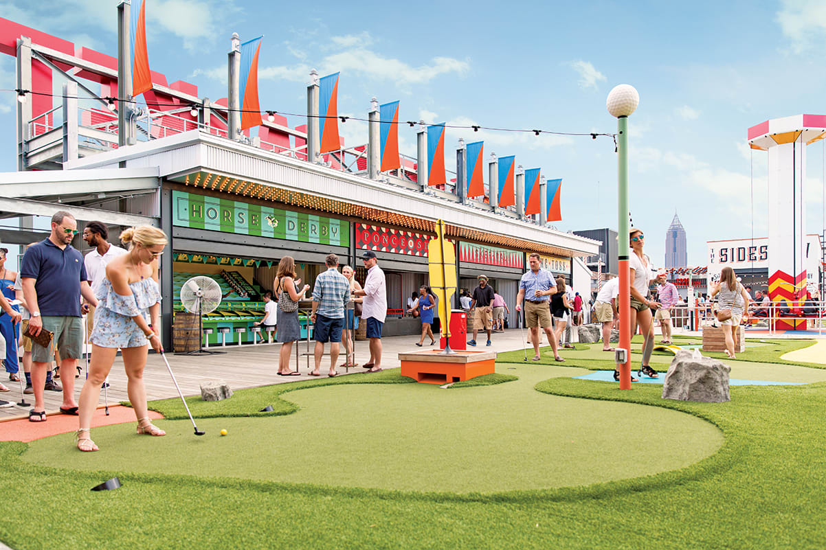 People play mini-golf on a rooftop course with colorful flags and buildings in the background in Atlanta, Georgia.