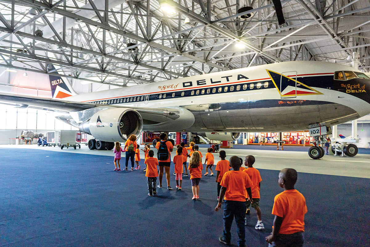 A group of children in orange shirts tour a large Delta airplane inside a hangar in Atlanta, Georgia.