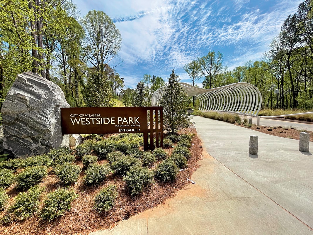 A winding path leads through a lush, green park with a distinctive white archway and a large rock sign in Atlanta, Georgia.