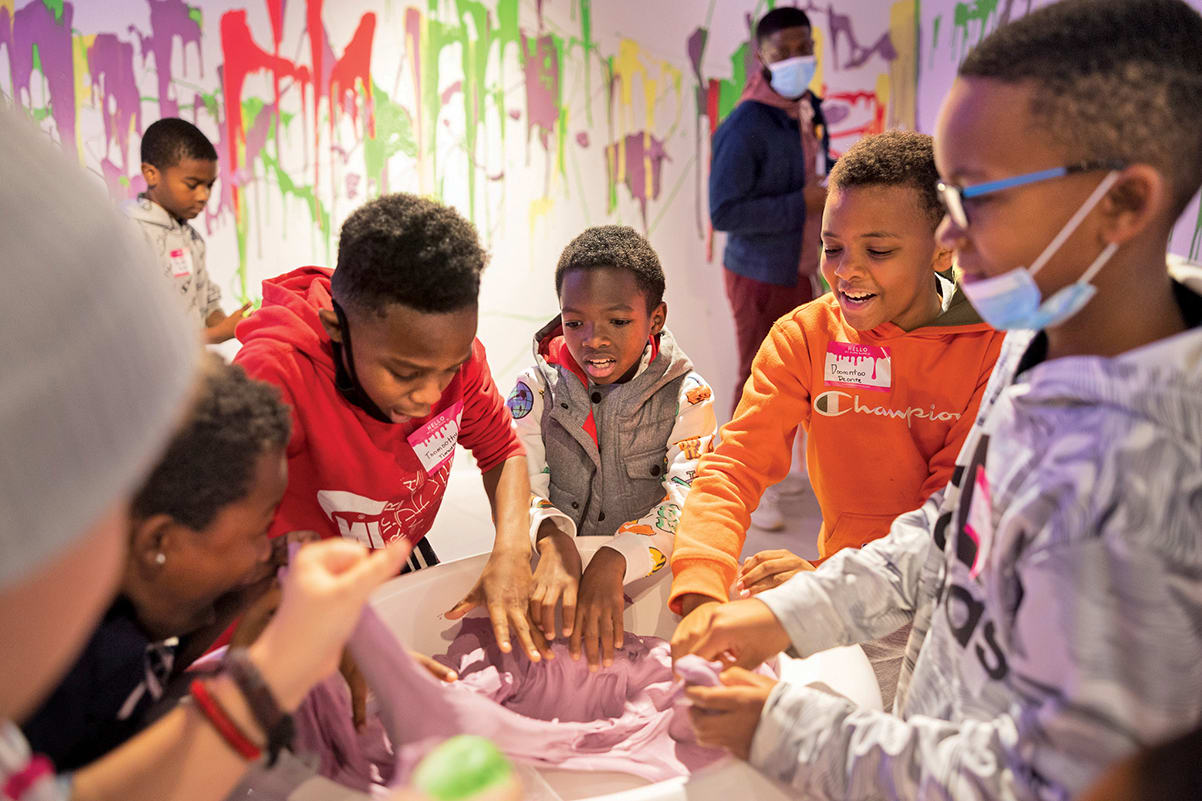 A group of children in colorful clothing are engaged in a hands-on activity, possibly making slime, in a room with vibrant graffiti-style wall art in Atlanta, Georgia.