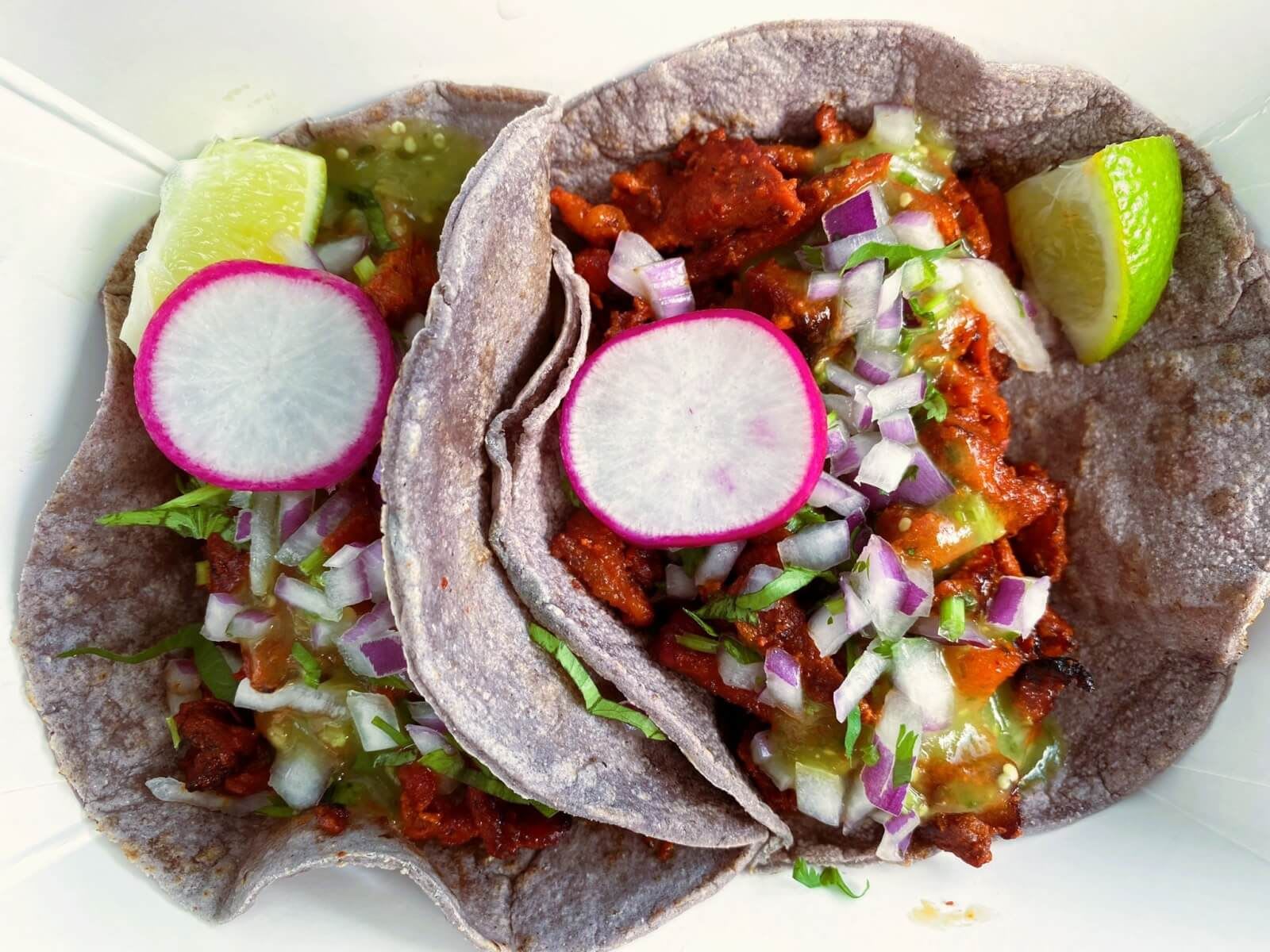 Two purple corn tacos filled with seasoned meat, onions, radishes, cilantro, and lime wedges in Atlanta, Georgia.