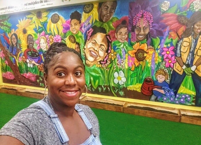 A woman smiles in front of a vibrant mural of diverse people among colorful flowers in Atlanta, Georgia.