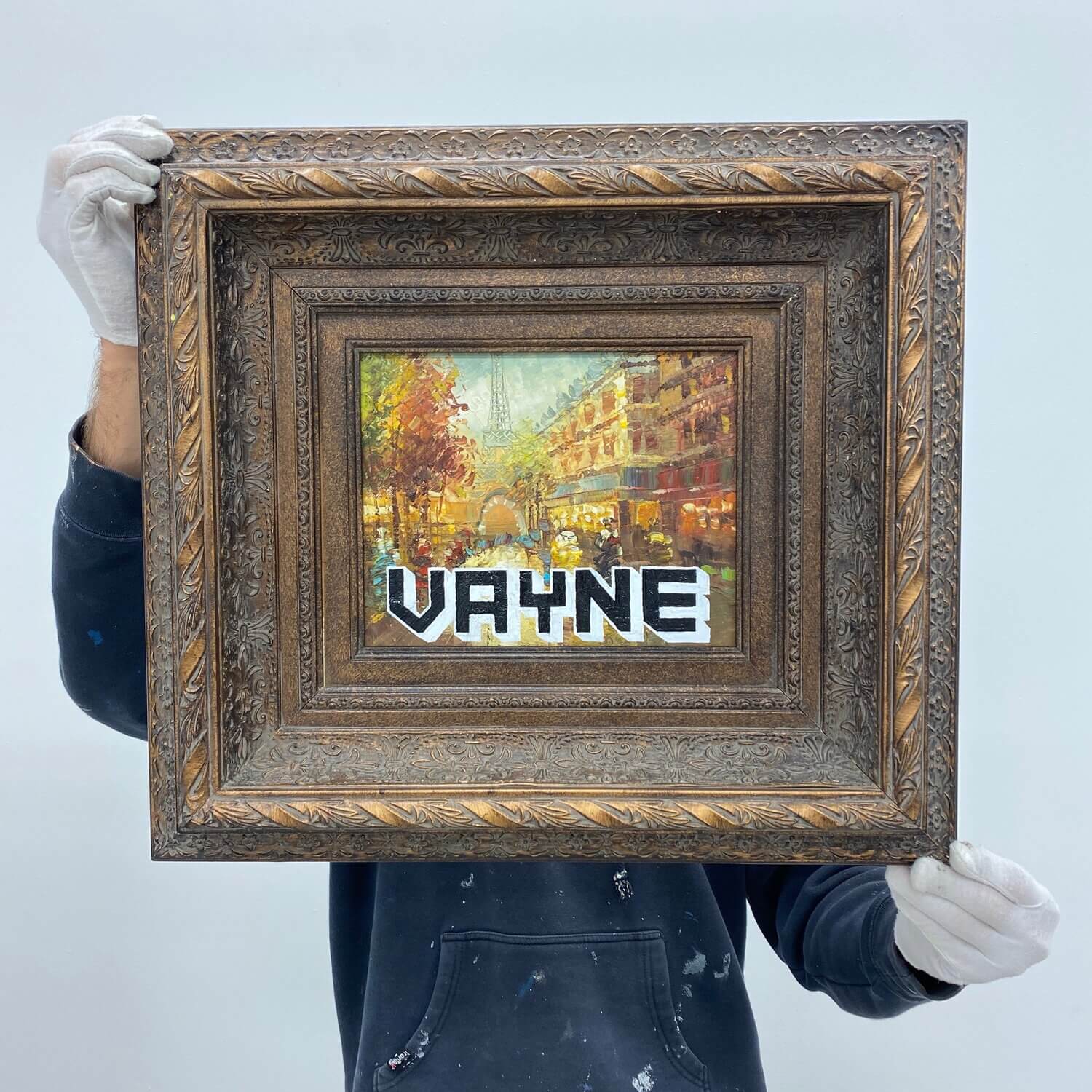 A person in a black hoodie holds a framed painting of a Parisian street scene with the word 'VAYNE' superimposed in Atlanta, Georgia.