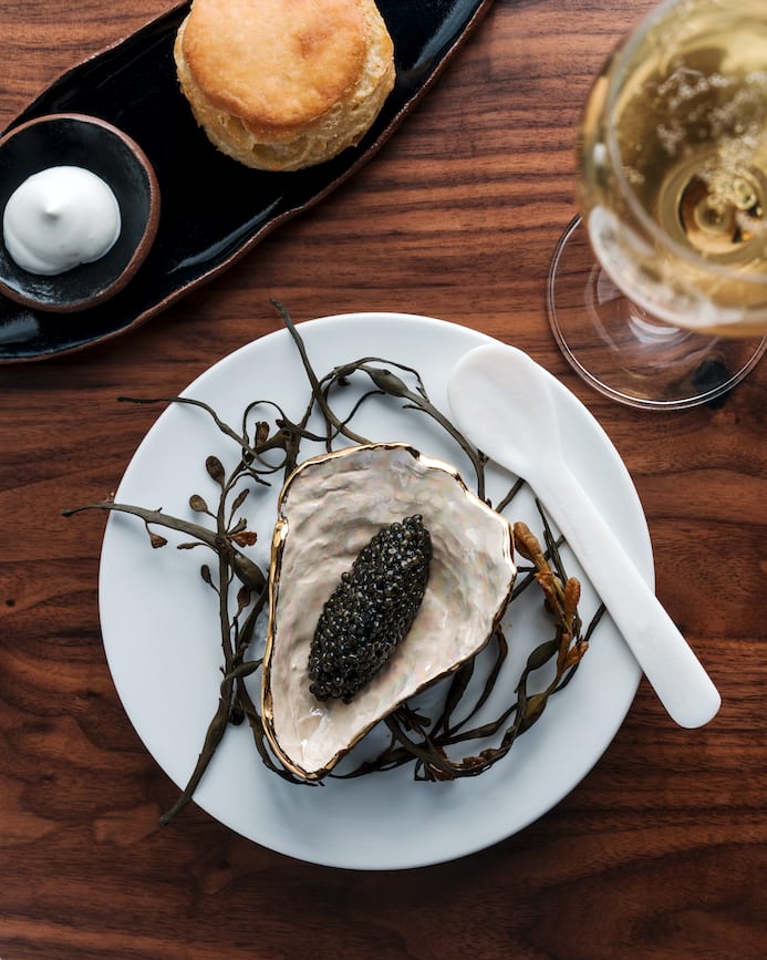 One Day Neighborhood Hopping Around Atlanta 9 A white plate on a wooden table holds a golden oyster shell filled with black caviar, surrounded by seaweed and accompanied by a white spoon, a glass of white wine, and a black plate with a biscuit and a small bowl of white sauce in Atlanta, Georgia.