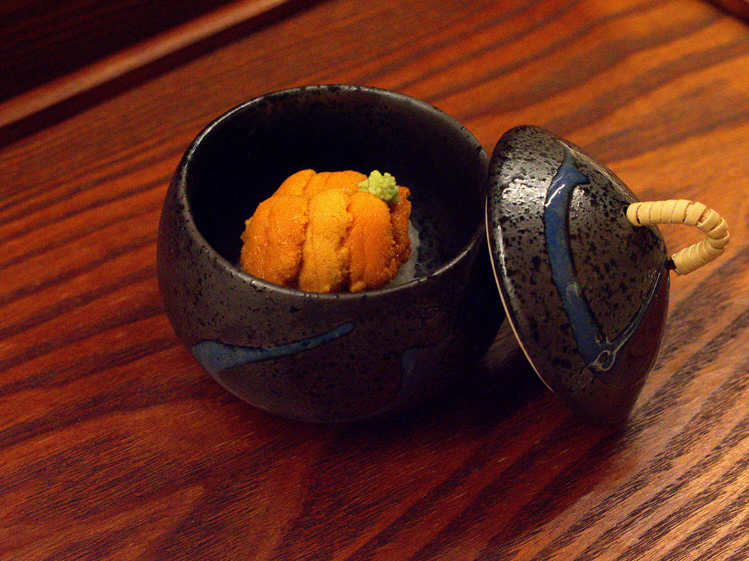 A small, dark ceramic bowl with a lid contains a piece of orange uni (sea urchin) with green wasabi on top in Atlanta, Georgia.