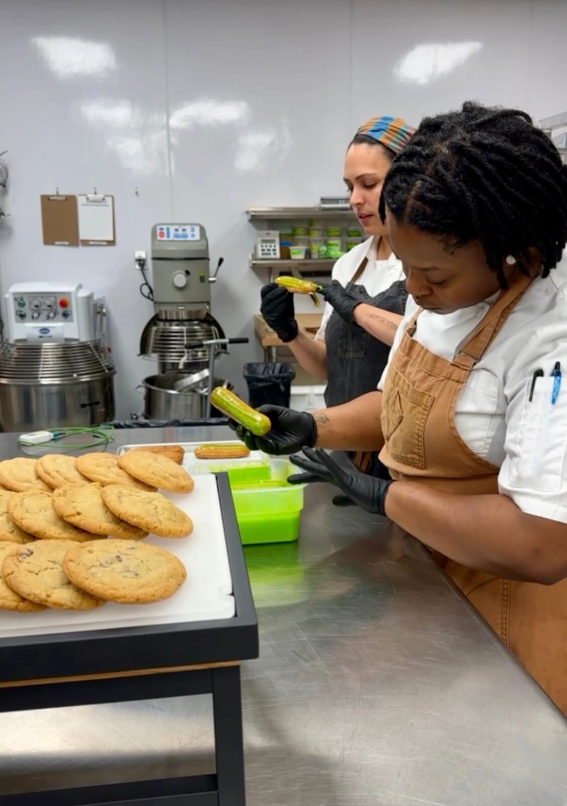 Two women in aprons and gloves decorate cookies in a professional kitchen in Atlanta, Georgia.