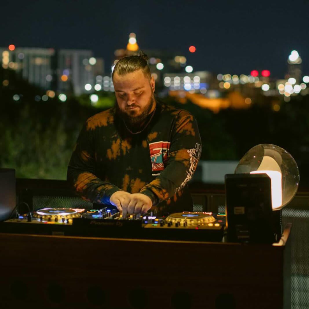 A DJ with a beard mixes music on a rooftop at night with city lights in the background in Atlanta, Georgia.