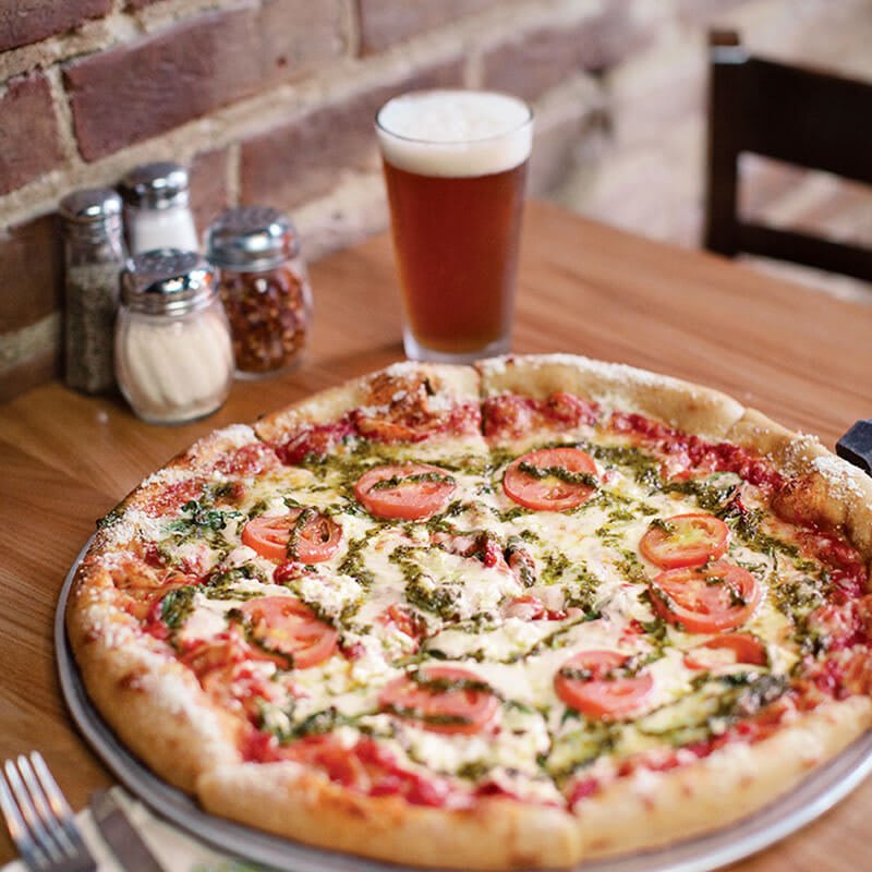 A large pizza with tomatoes, cheese, and pesto sauce sits on a wooden table with a glass of beer in Atlanta, Georgia.