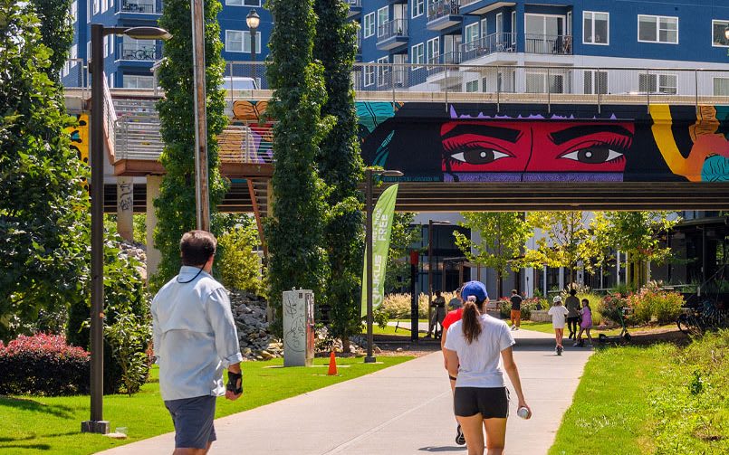 A vibrant urban park features a colorful mural of a woman's face on an overpass, with people walking and biking along a tree-lined path below in Atlanta, Georgia.