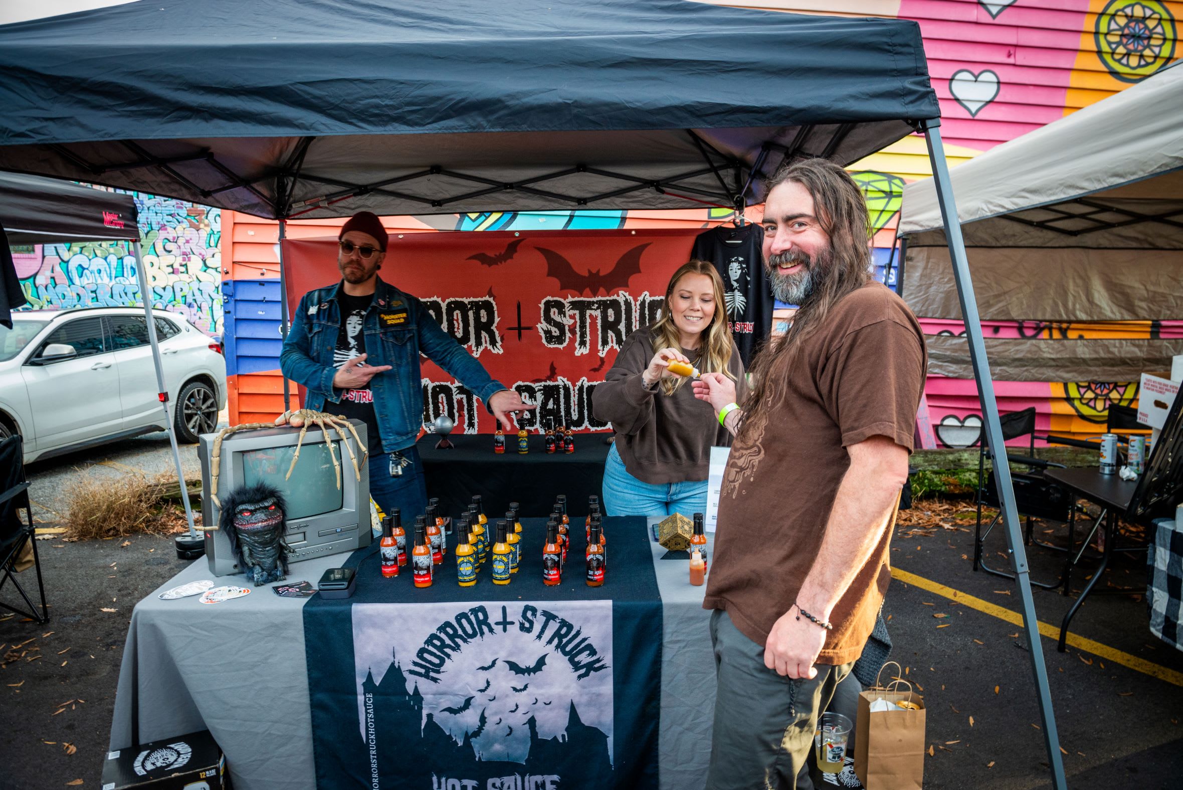 Three people sell hot sauce at an outdoor market under a black tent with Halloween decorations in Atlanta, Georgia.