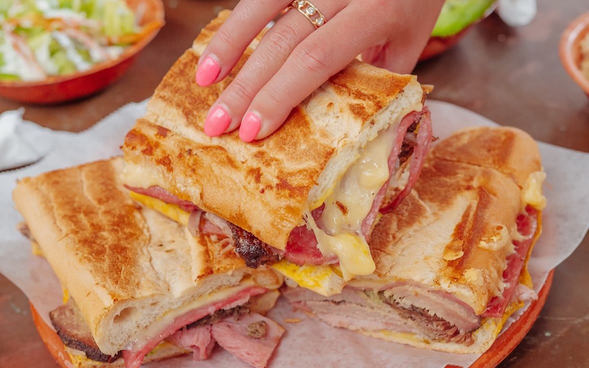 A hand with pink nail polish holds a toasted Cuban sandwich with melted cheese and meats in Atlanta, Georgia.