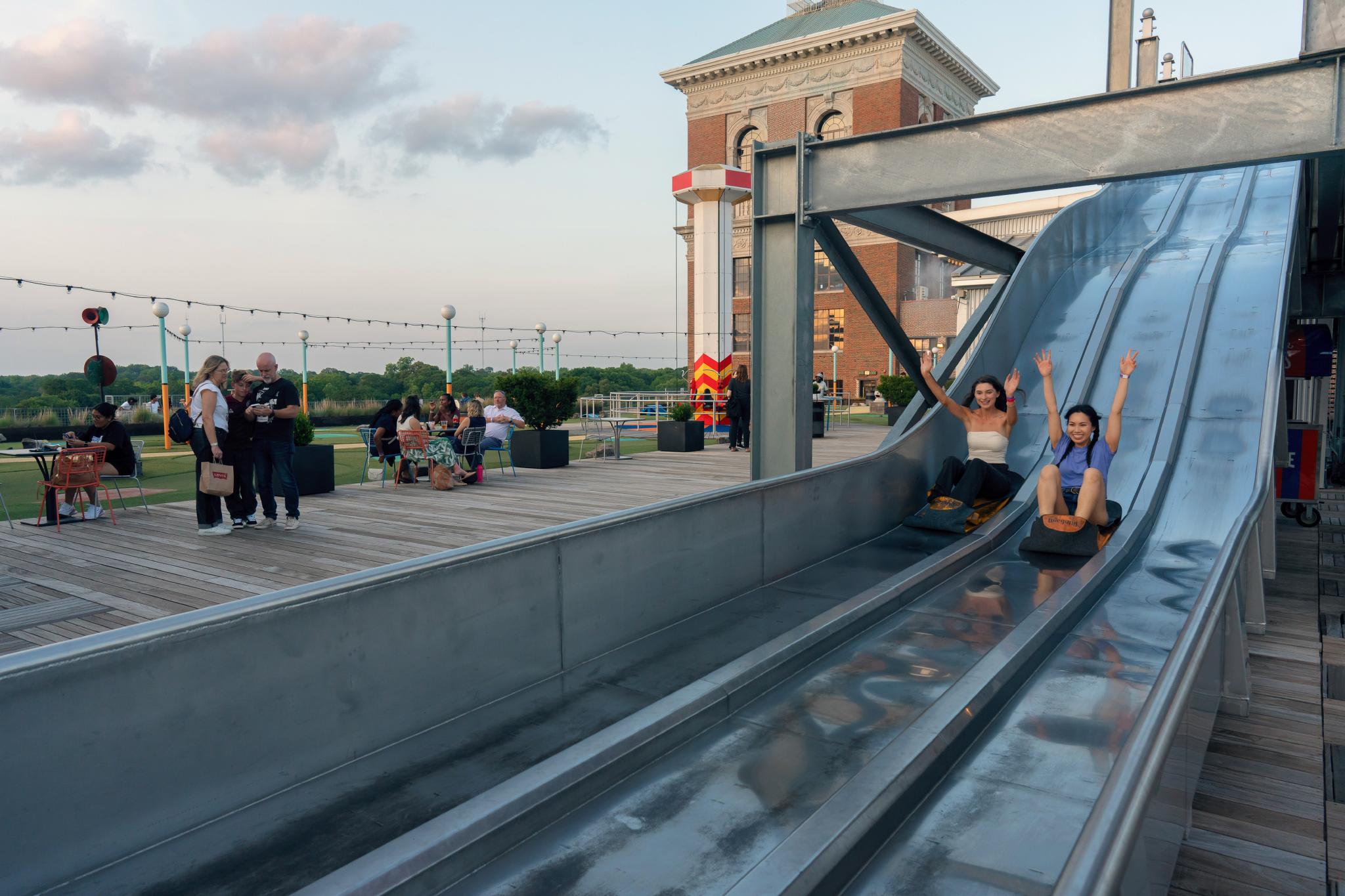 Two women slide down a long, metallic slide on a rooftop amusement park in Atlanta, Georgia.