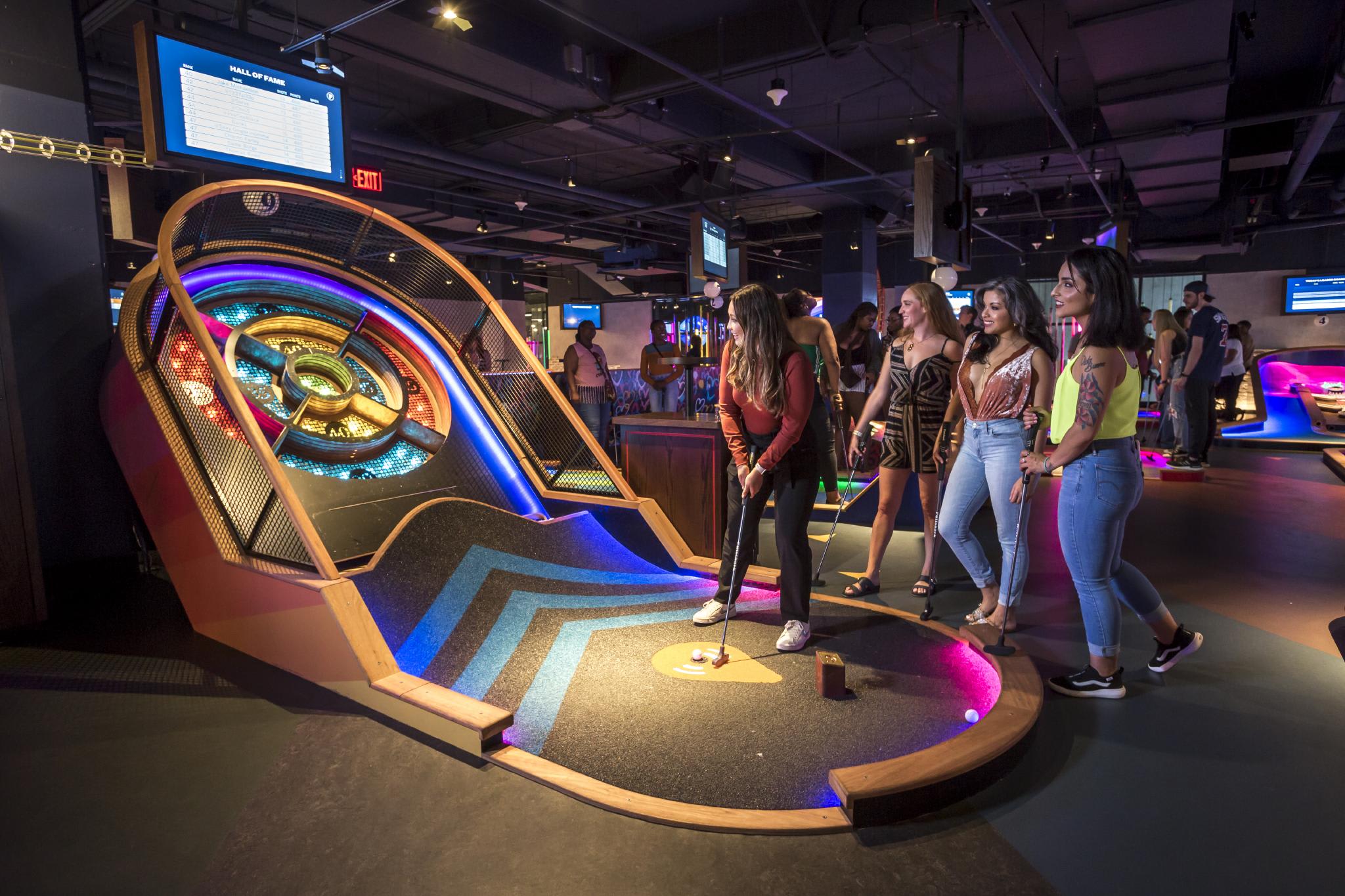 Women play mini-golf in a neon-lit indoor course with interactive scoreboards in Atlanta, Georgia.