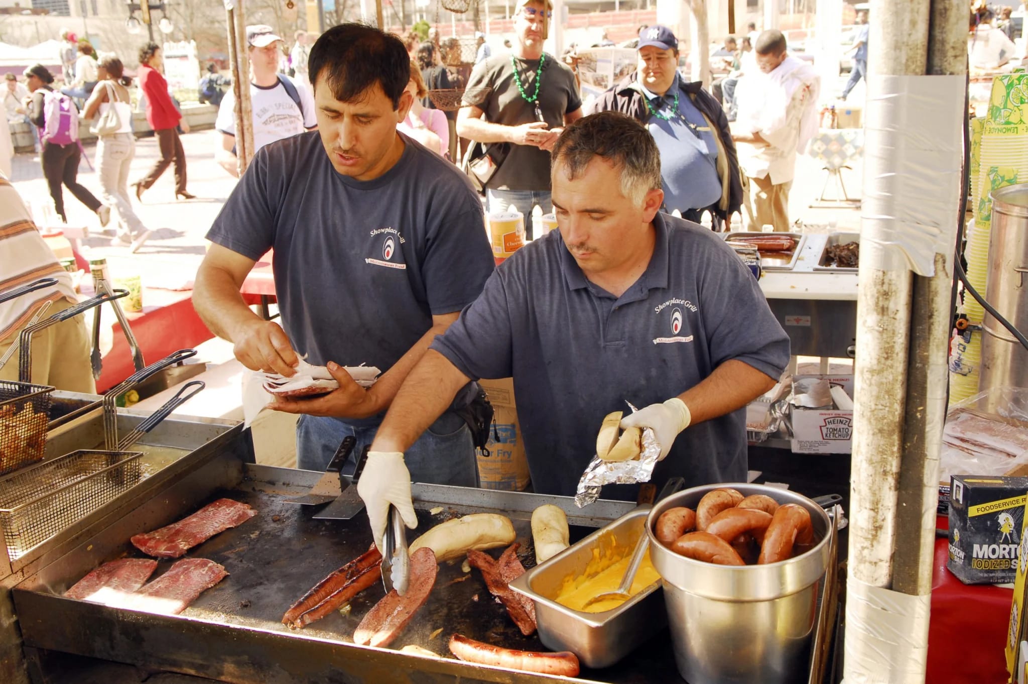 Two men in blue shirts cook hot dogs and sausages on a grill at a food stand in Atlanta, Georgia.