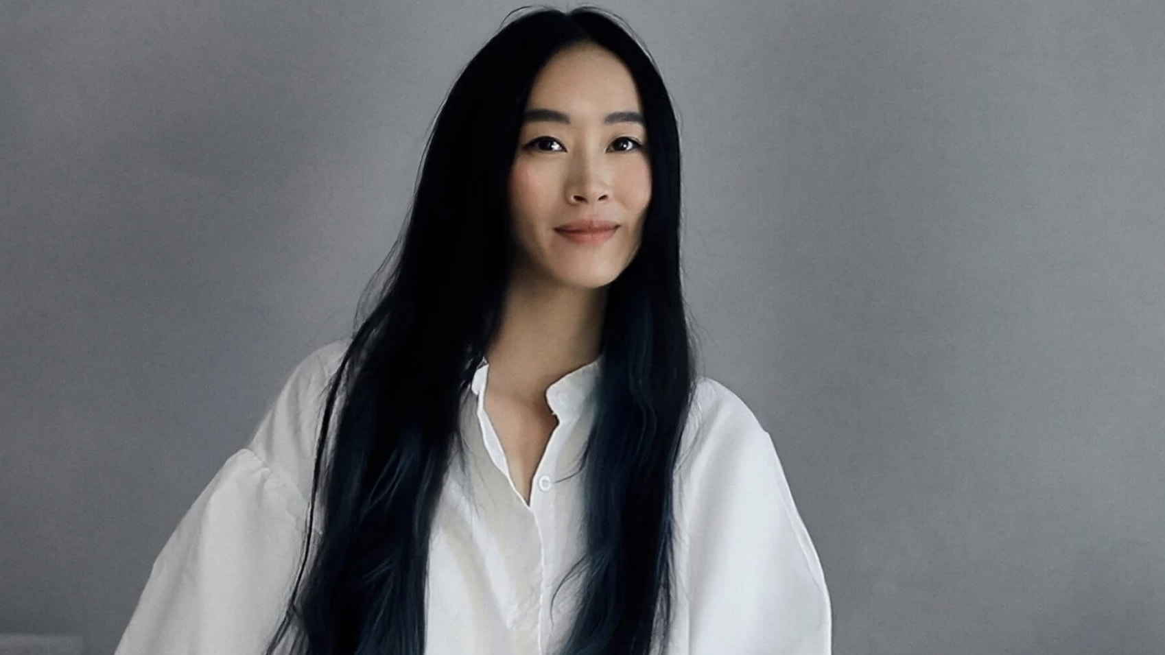 A woman with long, dark hair and a gentle smile stands against a gray wall in Atlanta, Georgia.