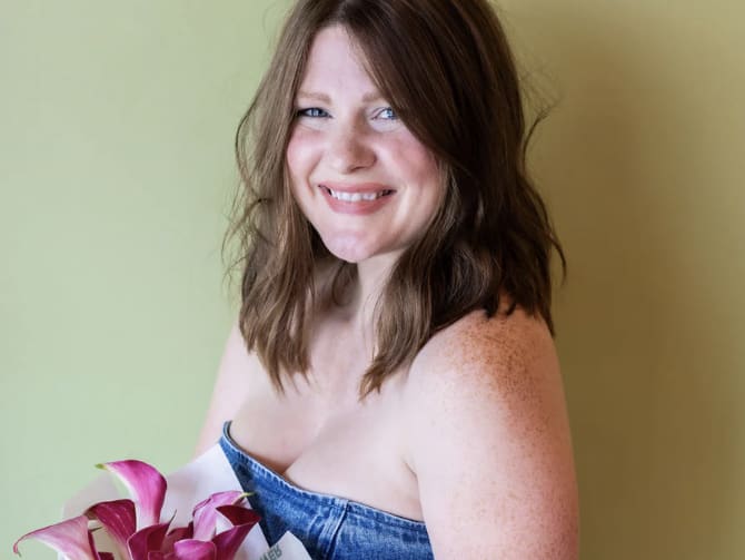 A smiling woman with freckles holds a bouquet of pink flowers against a green wall in Atlanta, Georgia.