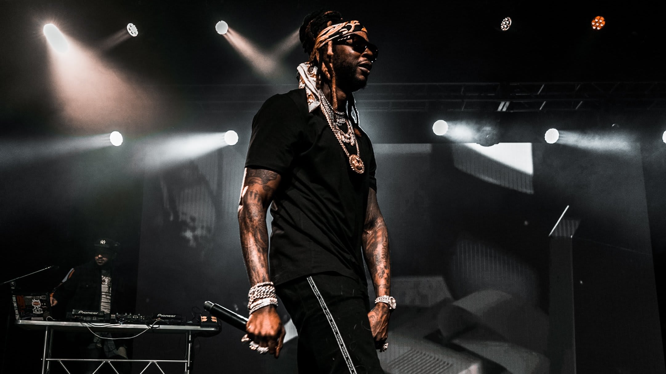 A musician performs on stage, wearing a black outfit and jewelry, with a DJ in the background in Atlanta, Georgia.