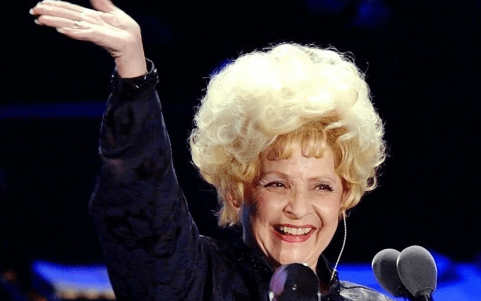 An elderly woman with a large, voluminous blonde wig is smiling and waving at an audience in Atlanta, Georgia.