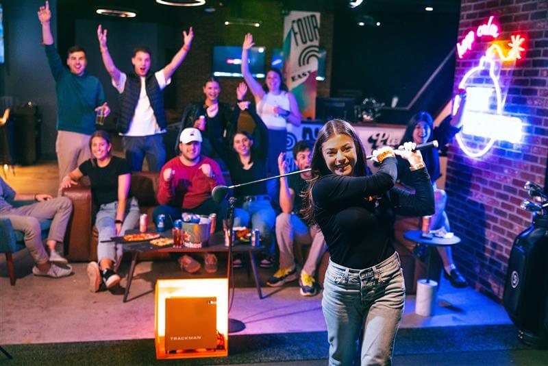 A woman golfs indoors while a group of friends cheers her on in a dimly lit room with neon signs in Atlanta, Georgia.