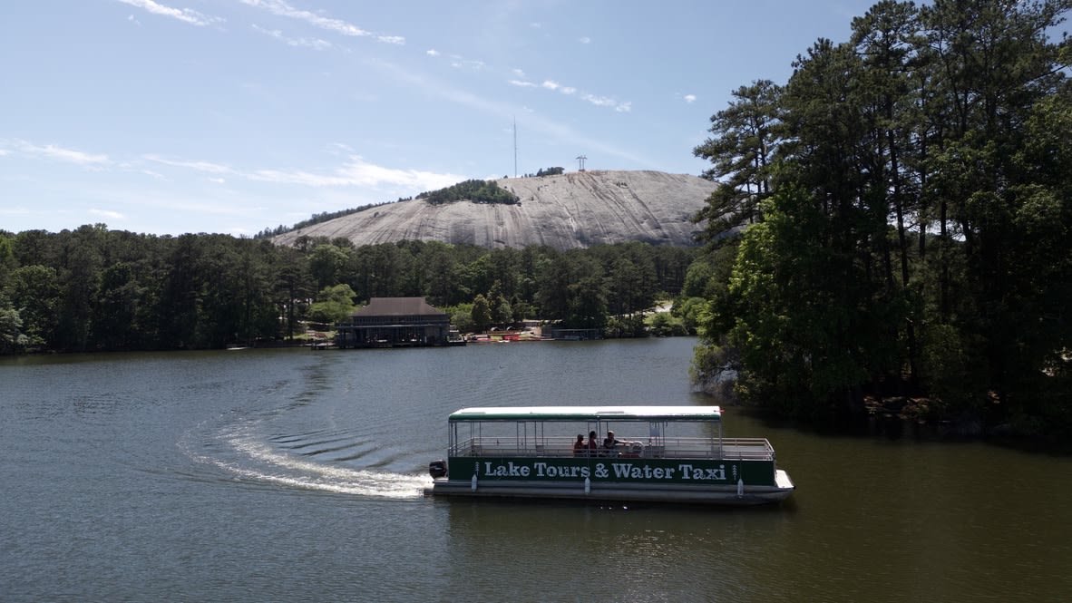 A green tour boat labeled 'Lake Tours & Water Taxi' cruises on a calm lake surrounded by trees in Atlanta, Georgia.
