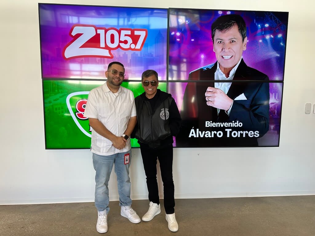 Two men stand in front of a colorful wall with radio station logos and a celebrity portrait in Atlanta, Georgia.