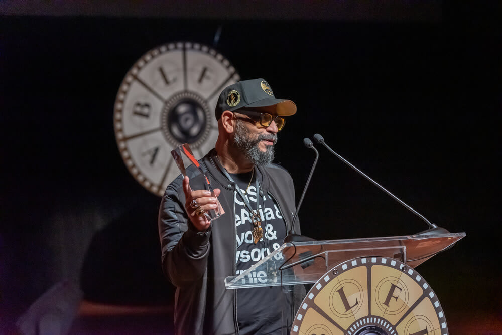 A man with a beard and glasses stands at a podium with a film reel design, holding an award in Atlanta, Georgia.