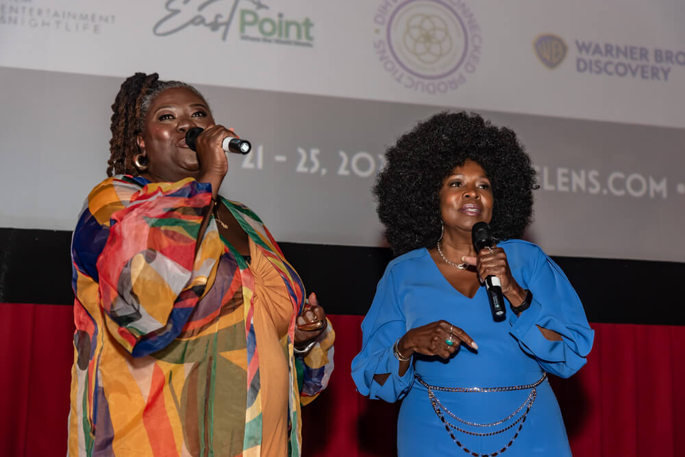 Two women stand on stage, one in a colorful patterned dress and the other in blue, both holding microphones in Atlanta, Georgia.