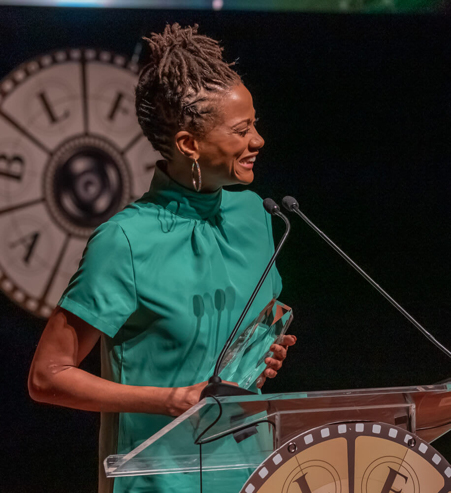BronzeLens Film Festival 5 A woman with dreadlocks, wearing a green dress, smiles while holding an award at a podium in Atlanta, Georgia.