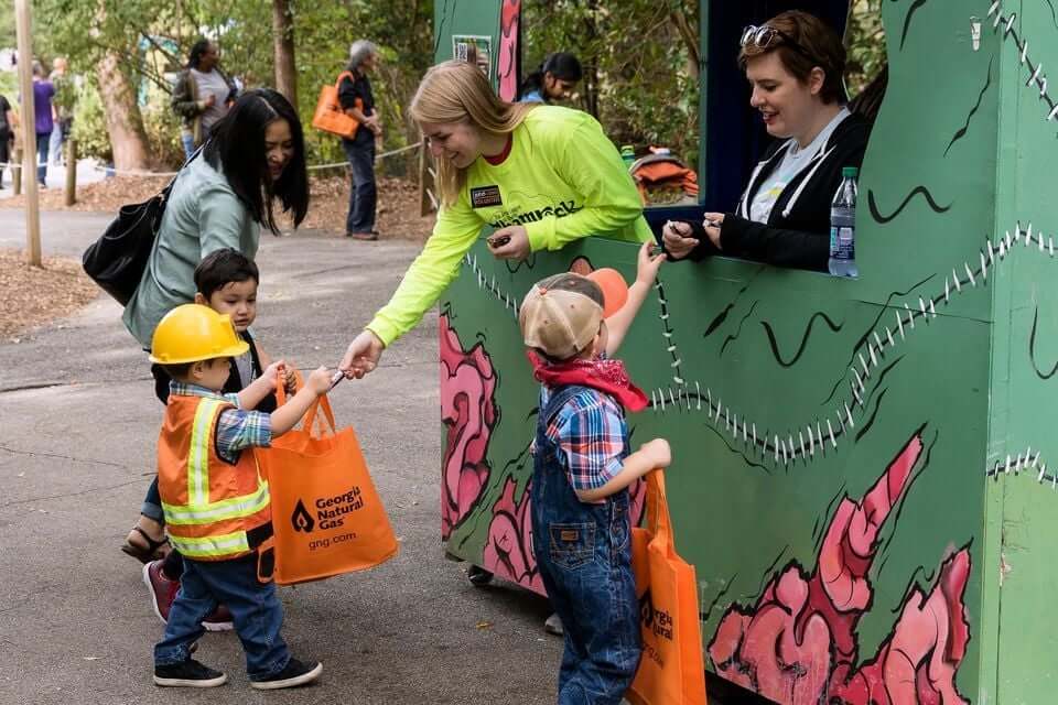 Children in construction gear receive orange bags from a woman at a colorful booth in Atlanta, Georgia.