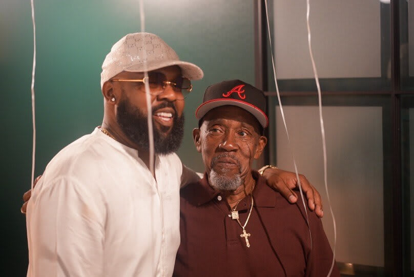 Two men, one young and one elderly, stand together smiling, dressed in casual attire in Atlanta, Georgia.