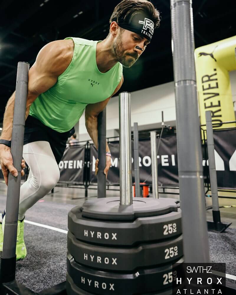Ultimate Guide to Hyrox Atlanta 4 Muscular man in green tank top and white tights lifts heavy weight plates on a gym machine in Atlanta, Georgia.