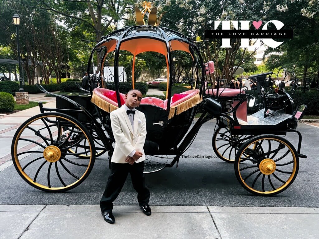 A young boy in a white tuxedo stands beside a black and gold horse-drawn carriage in Atlanta, Georgia.