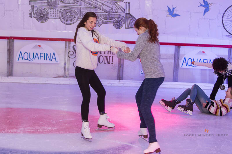 Two young women ice skating together, holding hands, with colorful lights on the ice rink in Atlanta, Georgia.