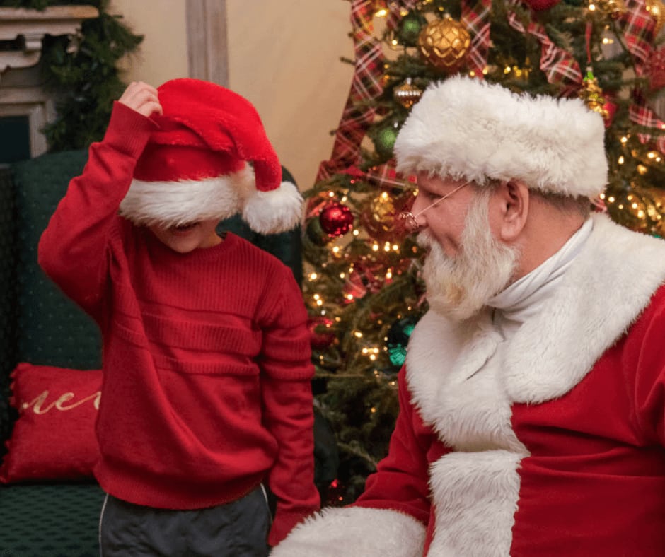 A young boy in a red sweater and Santa hat stands next to Santa Claus by a decorated Christmas tree in Atlanta, Georgia.