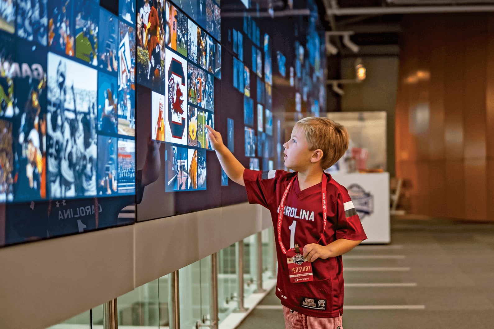 A young boy wearing a Carolina football jersey interacts with a large digital display wall in Atlanta, Georgia.