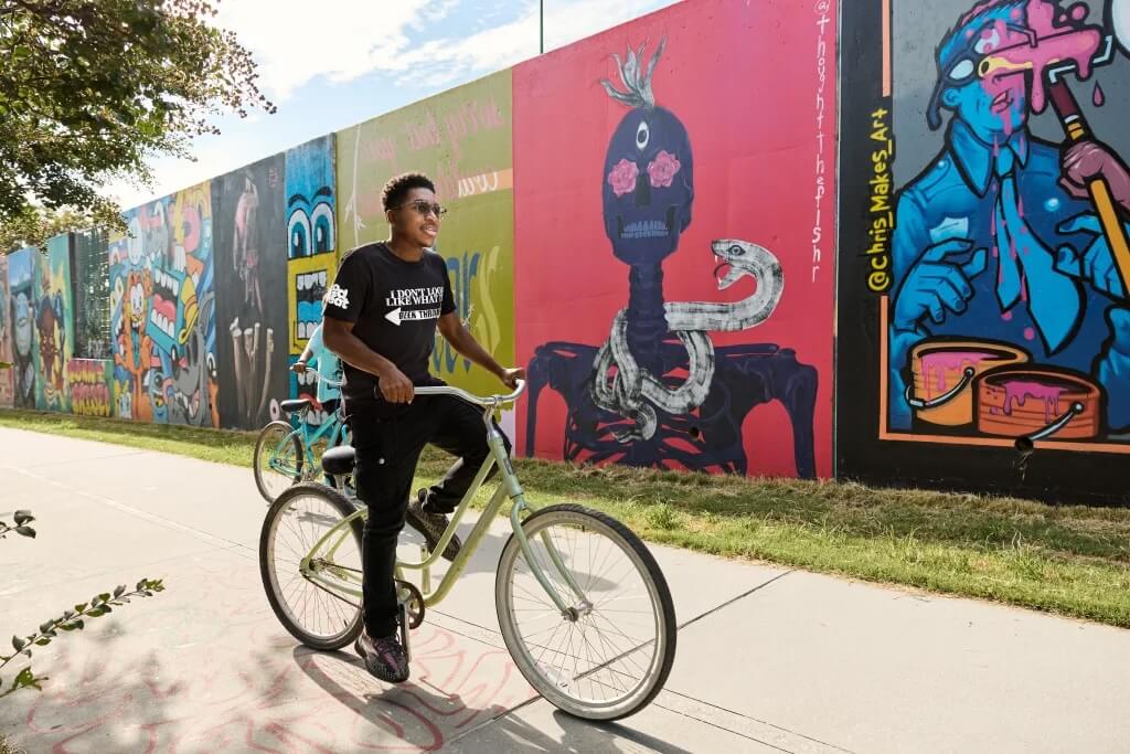 A young man rides a bicycle along a path with colorful graffiti murals on a wall behind him in Atlanta, Georgia.