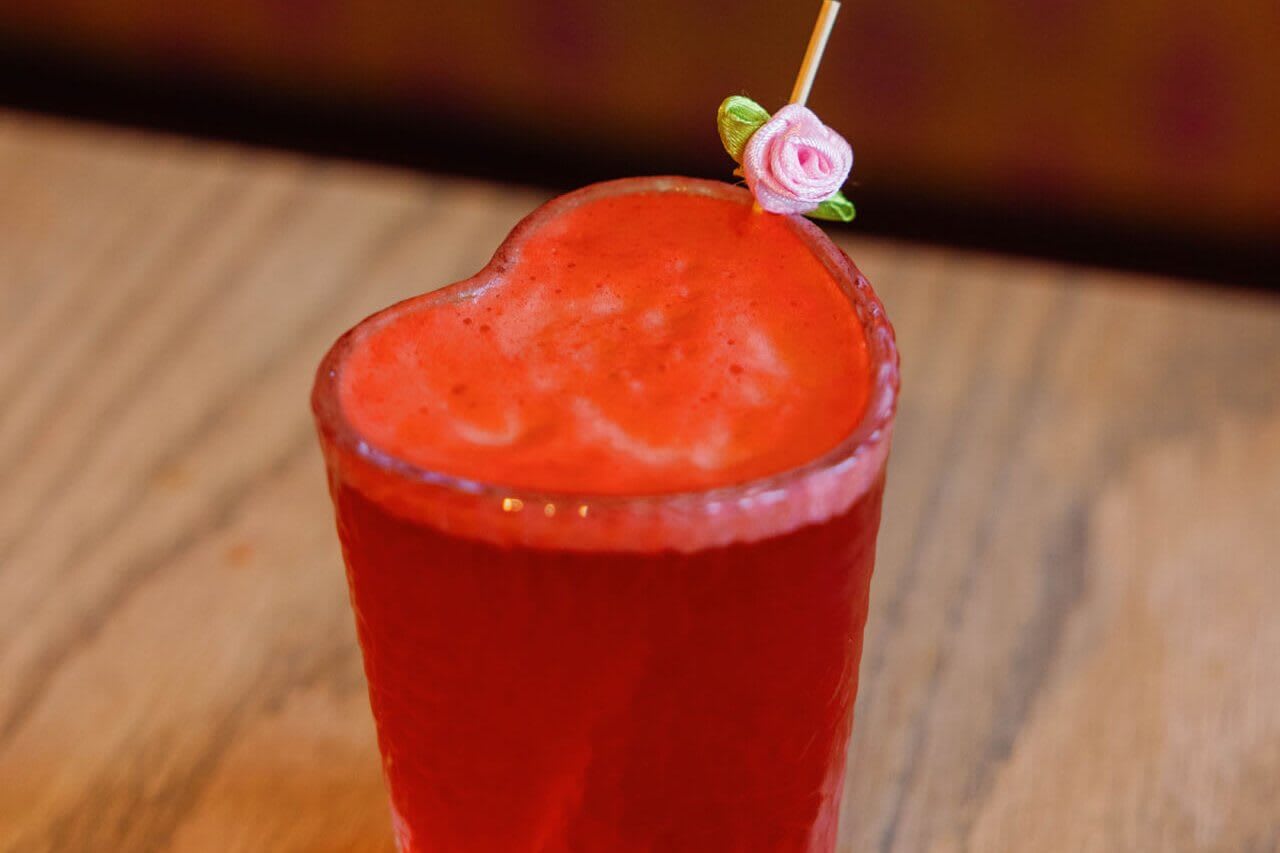 A vibrant red heart-shaped cocktail glass filled with a frothy beverage, garnished with a delicate pink rose and a straw, sits invitingly on a wooden table in Atlanta, Georgia.