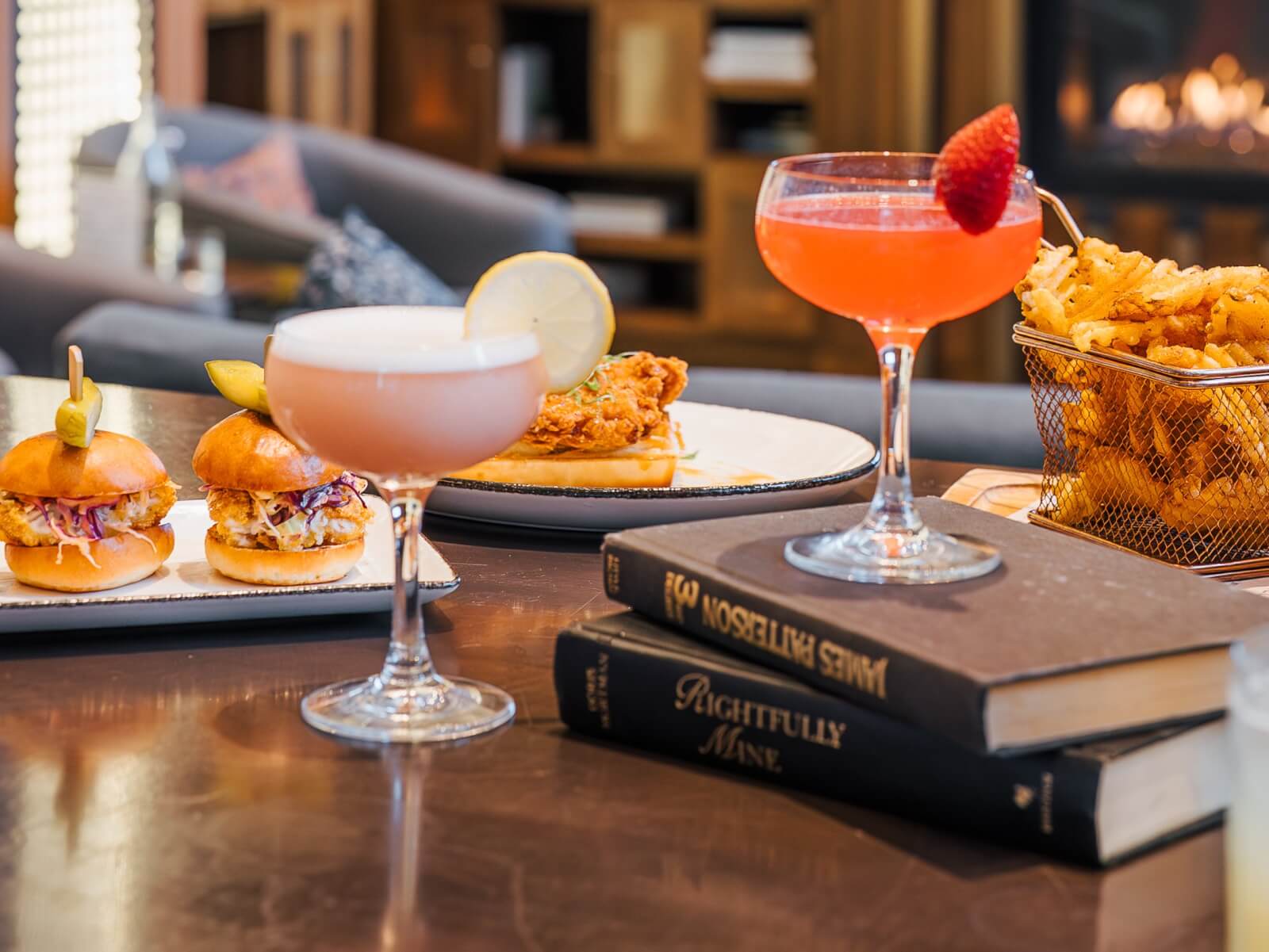 A cozy room with a fireplace features a table set with gourmet sliders, a variety of cocktails garnished with fruit, and a basket of crispy fries, all accompanied by a stack of books in Atlanta, Georgia.