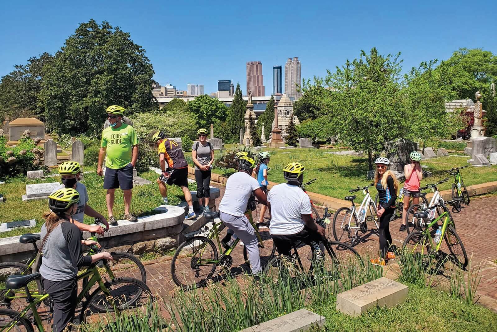 A group of cyclists wearing helmets and bright clothing ride through a cemetery with gravestones and trees in Atlanta, Georgia.